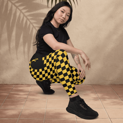 Person wearing a black t-shirt and yellow checkered pants posing against a beige wall with a palm leaf.