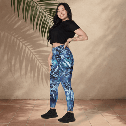 Woman wearing blue tie-dye leggings and black crop top standing against a beige wall with a palm leaf.