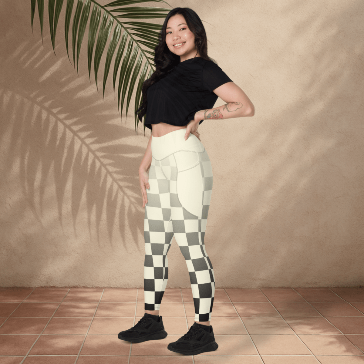 Woman wearing a black crop top and checkered leggings standing against a beige wall with a palm leaf.