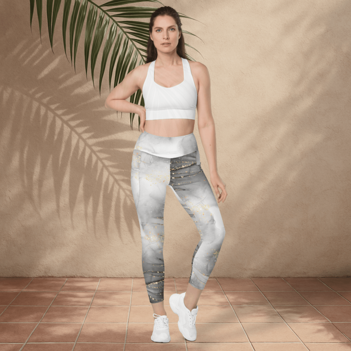 Woman wearing a white sports bra and patterned leggings standing against a beige wall with a plant.