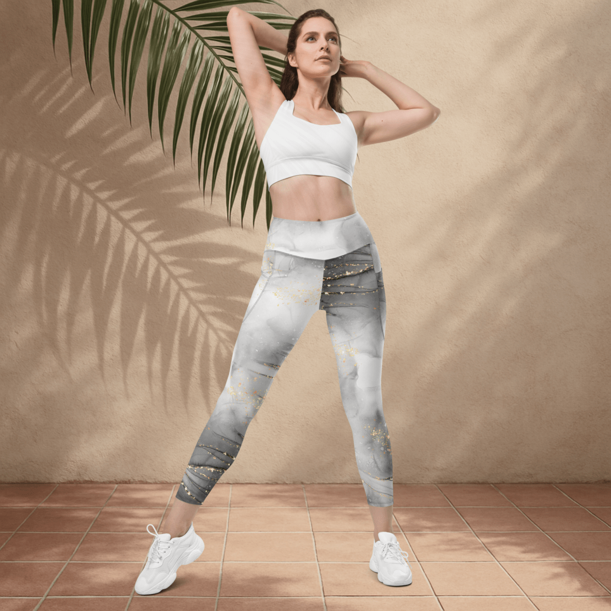 Woman wearing a white sports bra and marble leggings standing against a beige wall with a palm leaf.