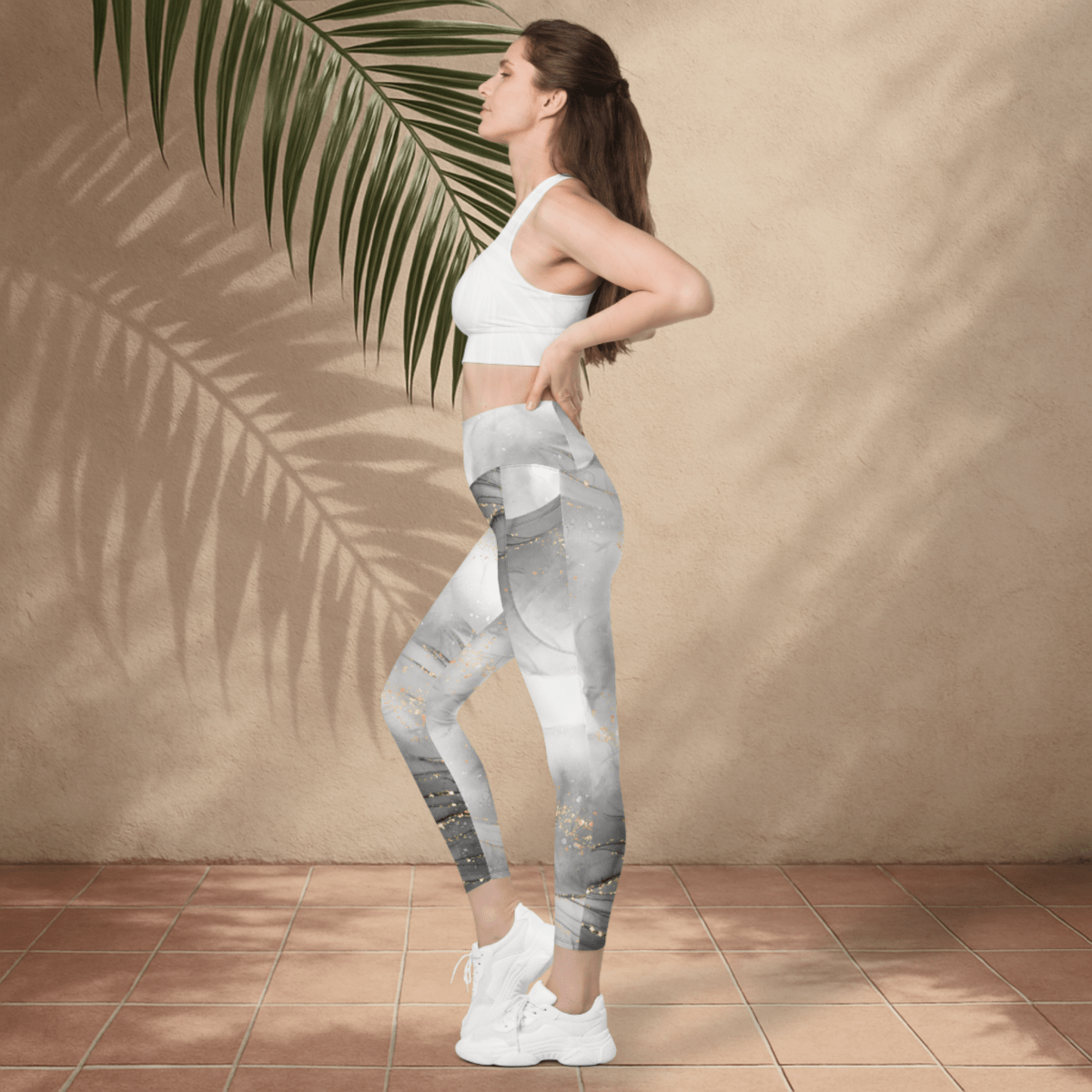 Woman wearing a white sports bra and gray leggings standing against a beige wall with a palm leaf.