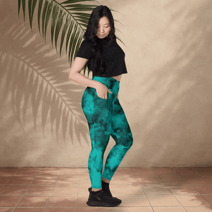 Woman wearing teal pants and black top standing against a beige wall with a leafy branch.