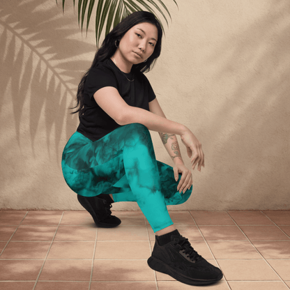 Woman in black t-shirt and teal leggings squatting on a tiled floor with a beige wall and plant in the background