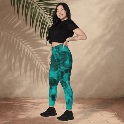 Woman wearing teal patterned leggings and black top standing against a beige wall with a plant leaf.