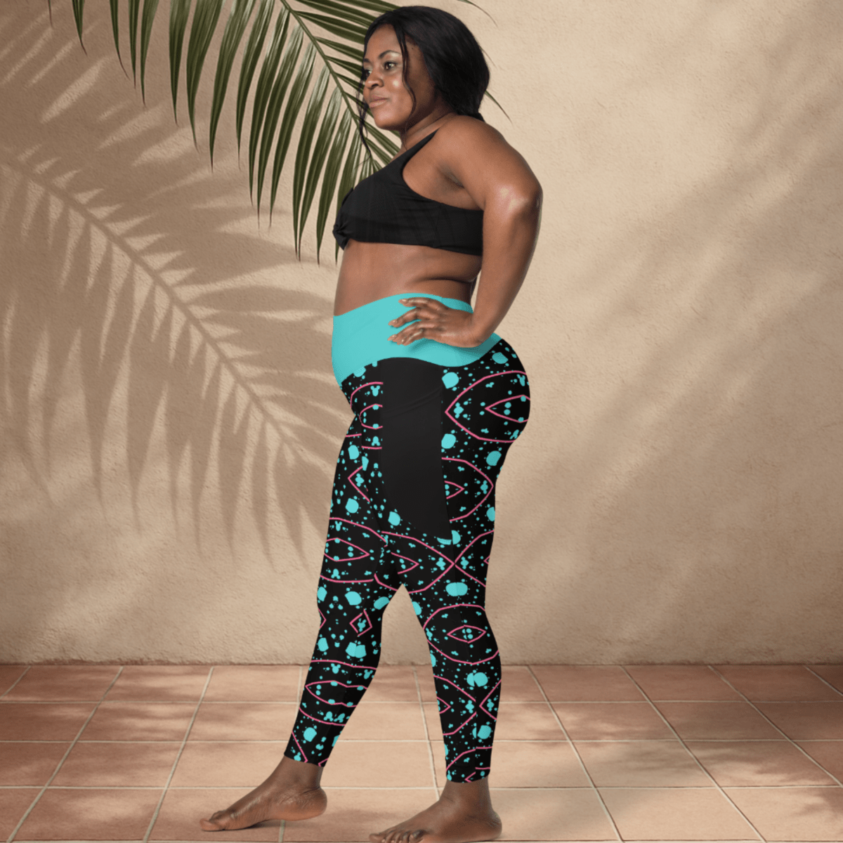 Woman wearing black and teal patterned leggings with a black sports bra against a beige wall.