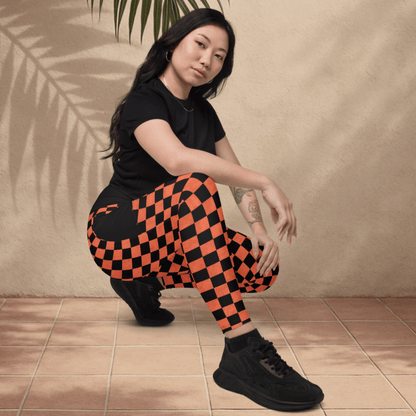 Person wearing black t-shirt and orange checkered pants posing against a beige wall with a palm leaf.