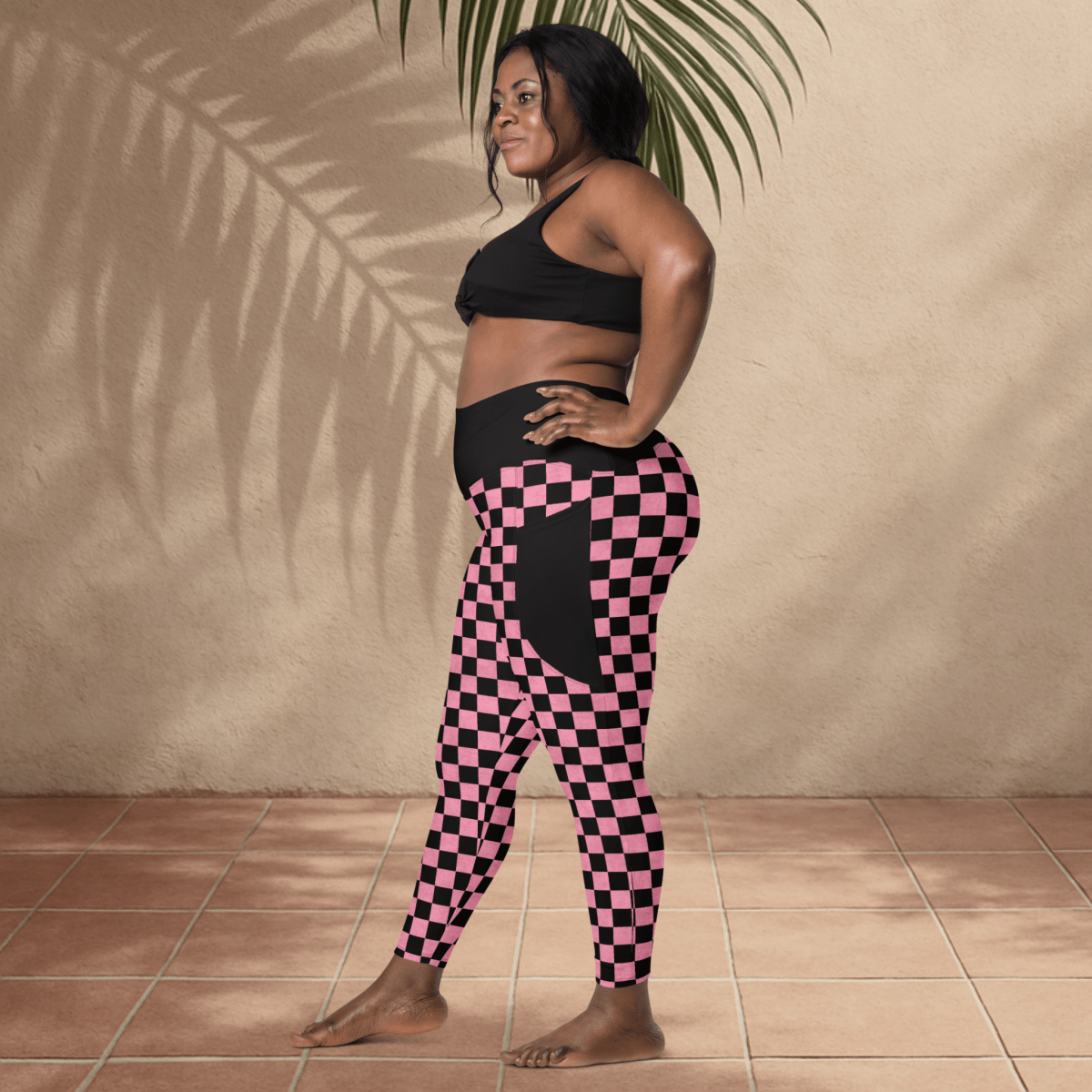 Woman wearing black sports bra and pink checkered leggings standing against a beige wall.