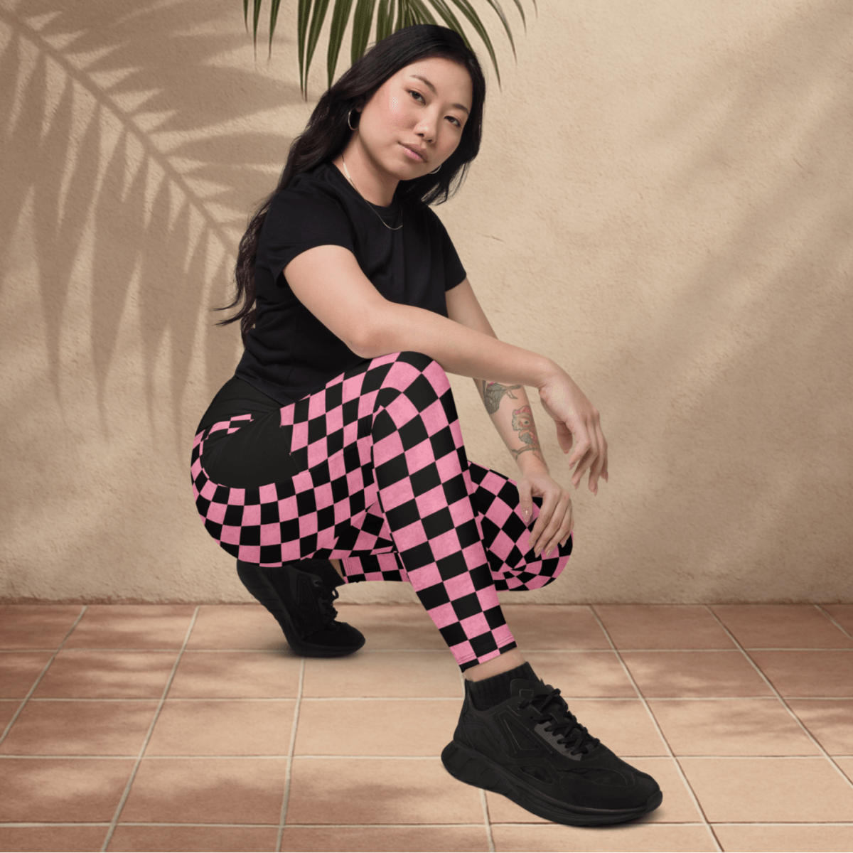 Person wearing a black t-shirt and pink checkered pants sitting against a beige wall with a palm leaf.
