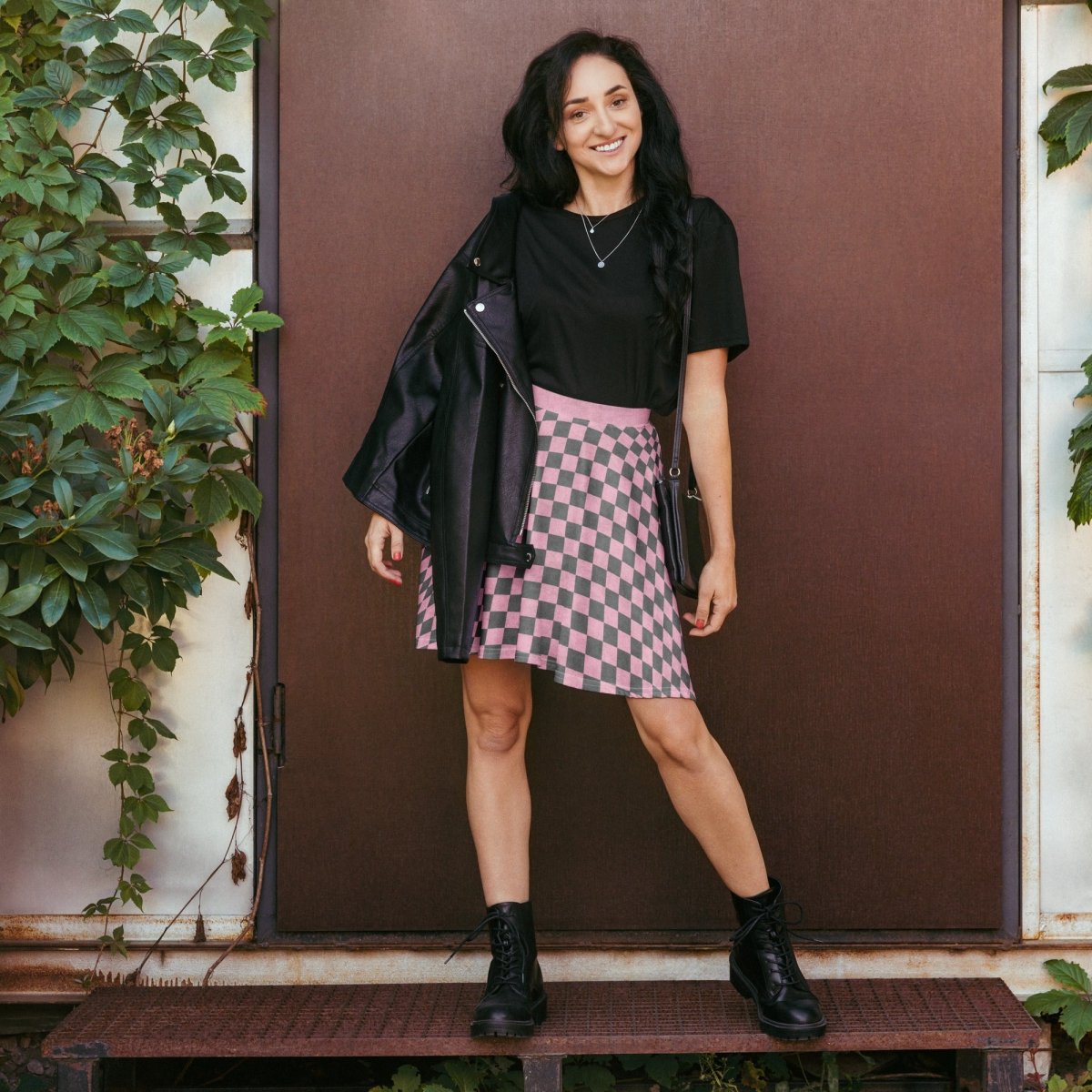 Woman in a black top and pink checkered skirt standing in front of a brown door.
