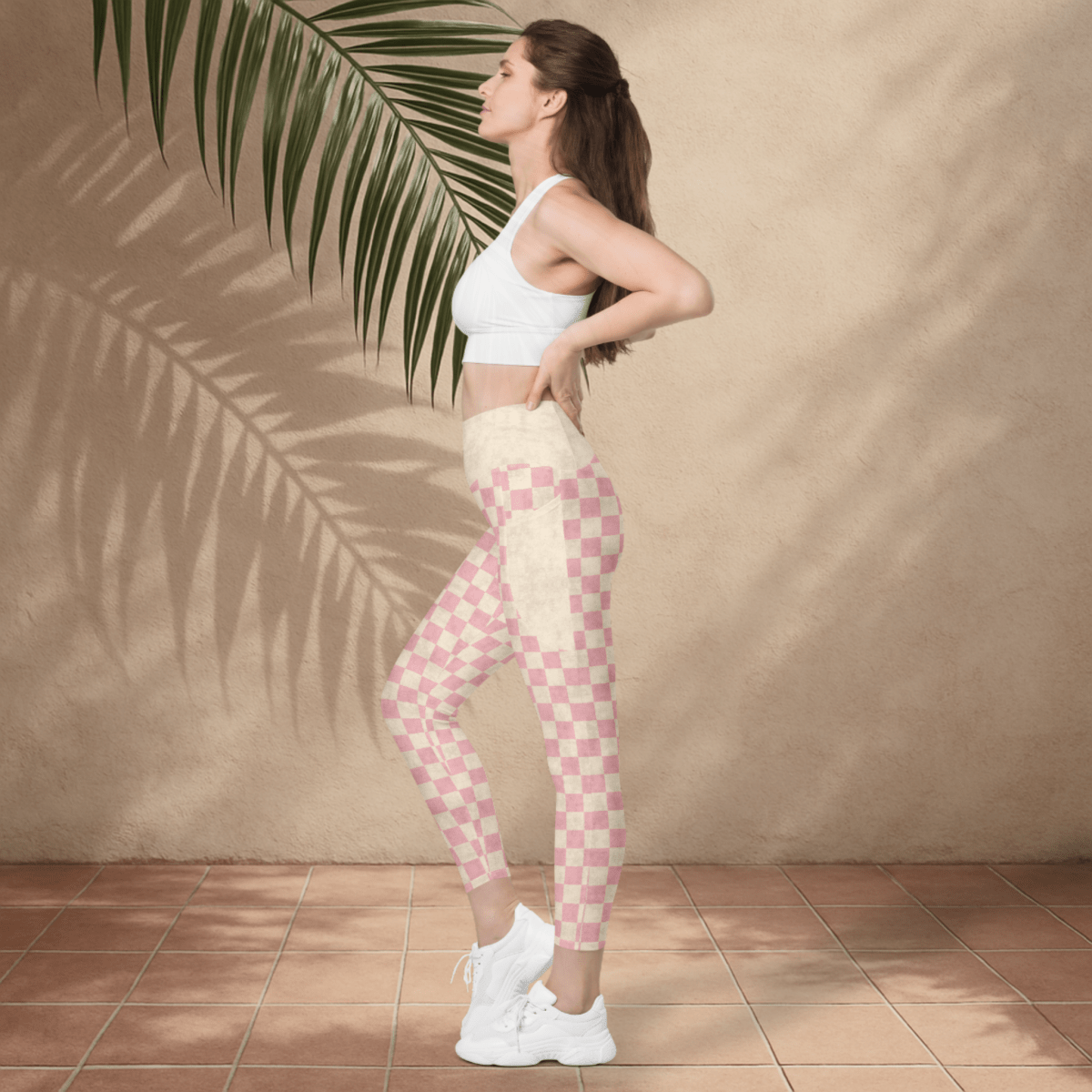 Woman wearing a white sports bra and pink checkered leggings standing against a beige wall with a palm leaf.