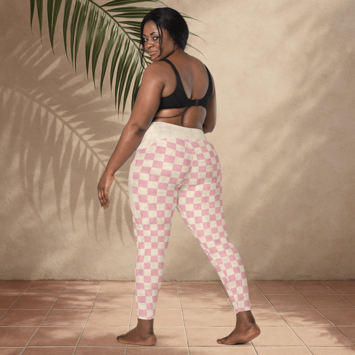 Woman wearing pink checkered leggings and black sports bra against a beige wall.