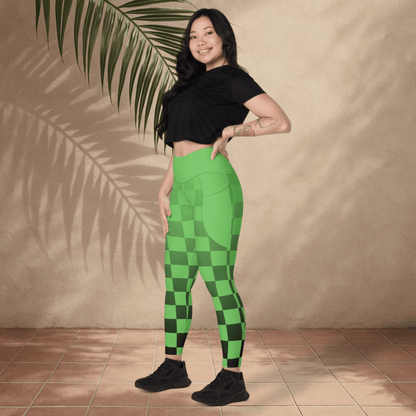 Woman wearing green checkered leggings and black top standing against a beige wall with a plant.