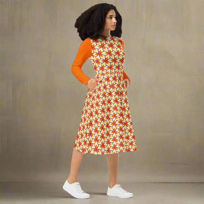 Slim, curly-haired model shown at a slight angle with both hands tucked into the side pockets of a bold triangle-patterned midi dress. The fitted bodice and flared skirt highlight the dress’s flattering silhouette. The long sleeves are a solid bright orange, contrasting with the geometric print in orange and cream. Set against a neutral gray background.