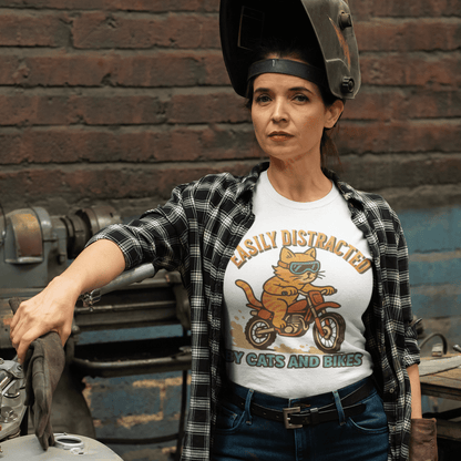 Woman wearing a graphic t-shirt with a cat on a motorcycle, standing next to a motorcycle in an industrial setting.