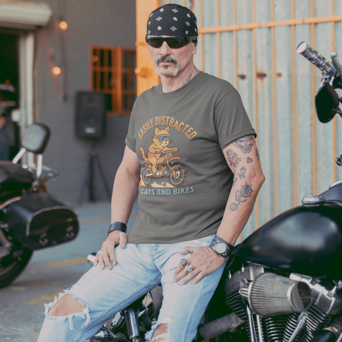 Man sitting on a motorcycle wearing a cat riding dirt bike graphic t-shirt and sunglasses, with another motorcycle in the background.