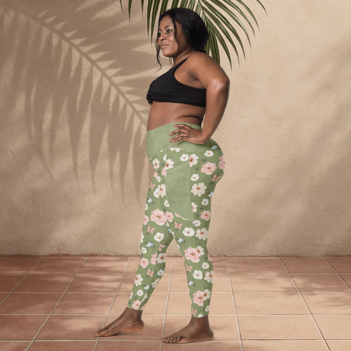 Woman wearing green floral leggings and black sports bra standing against a beige wall.