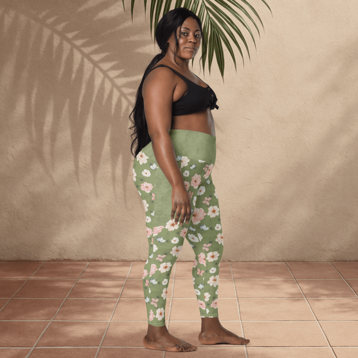 Woman wearing green floral leggings and black top standing against a beige wall.