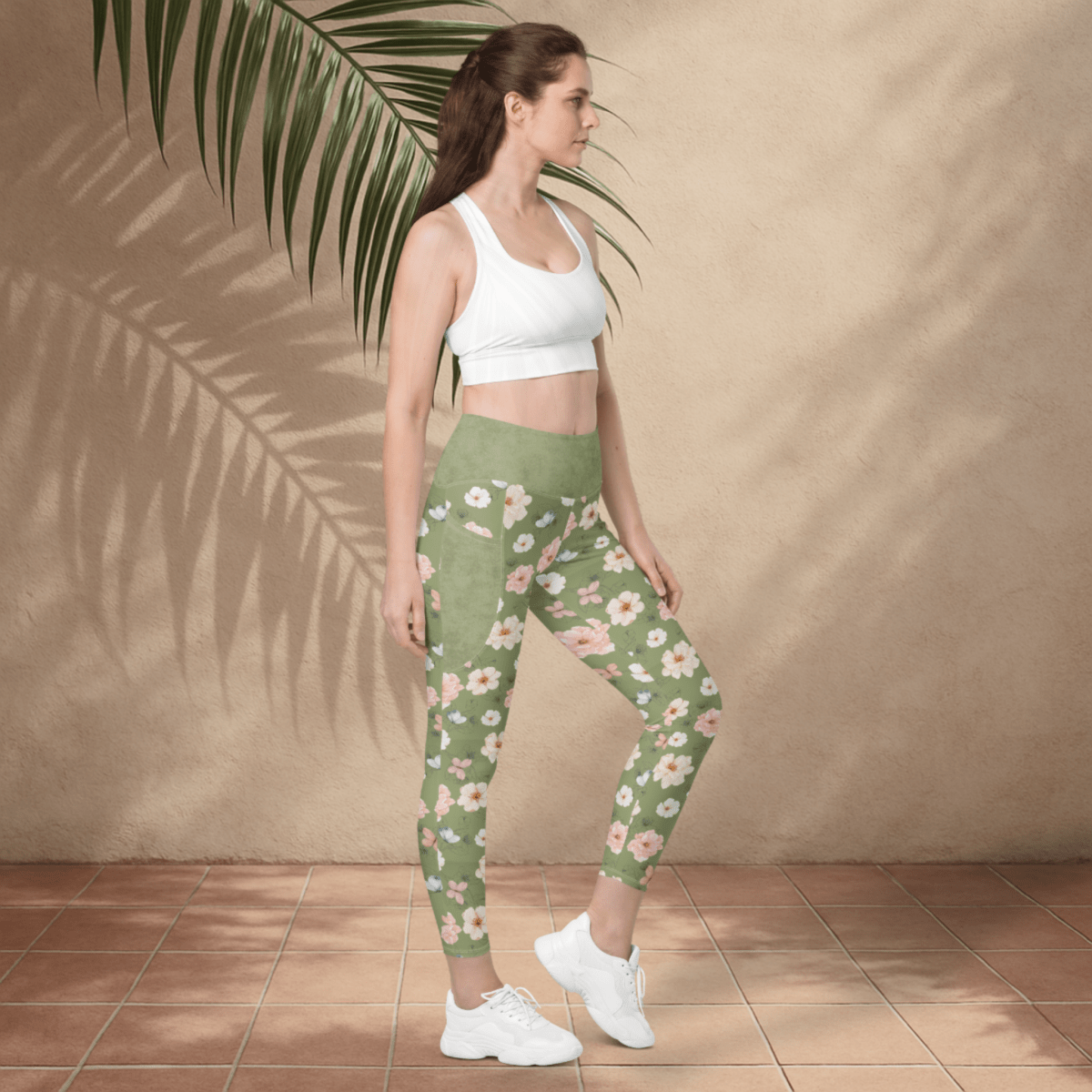 Woman wearing green floral leggings and white sports bra against a beige wall.