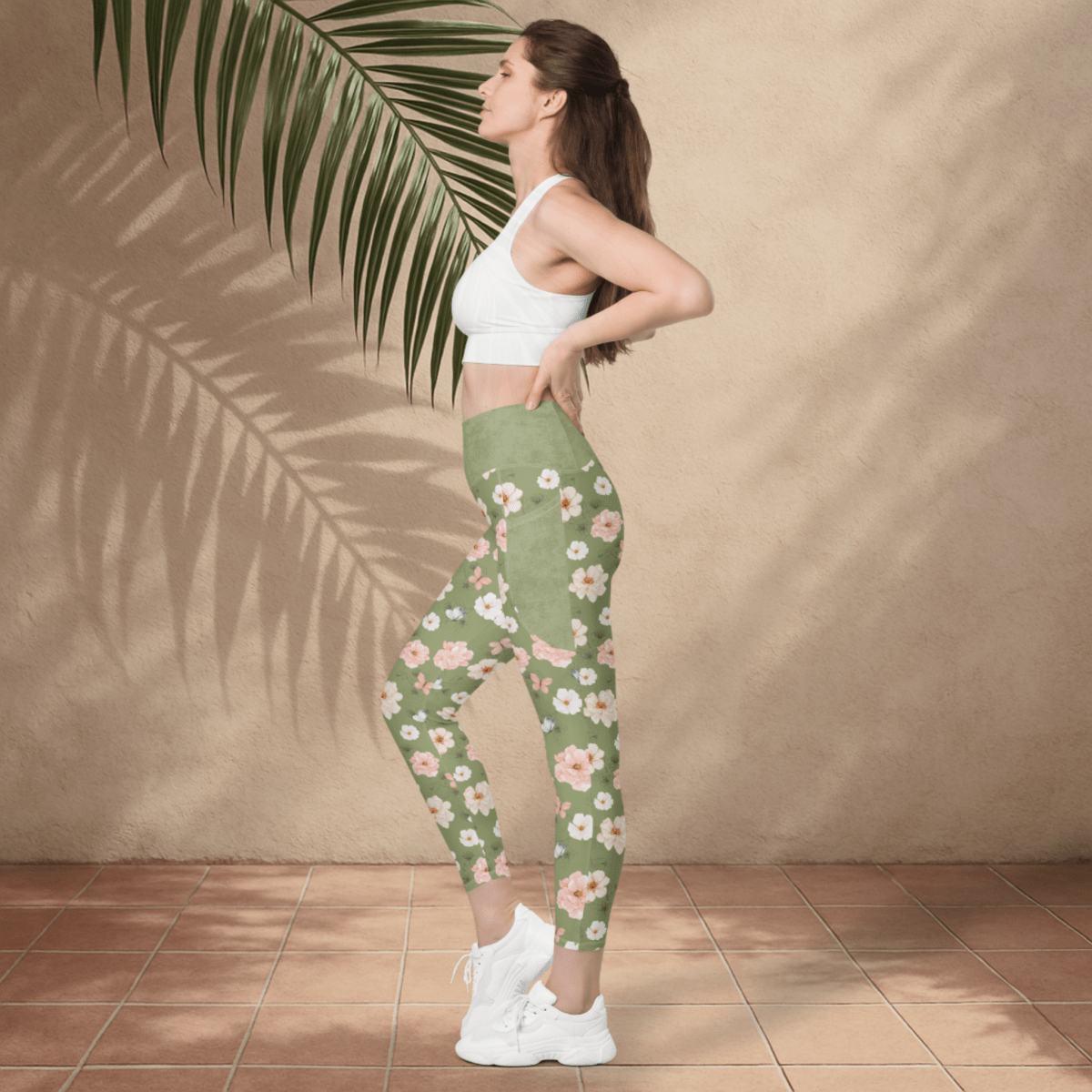 Woman wearing a white sports bra and green floral leggings standing against a beige wall with a palm leaf.