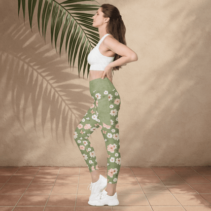 Woman wearing a white sports bra and green floral leggings standing against a beige wall with a palm leaf.