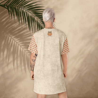 Person wearing a beige dress with a pumpkin design, standing against a beige wall with a palm leaf.
