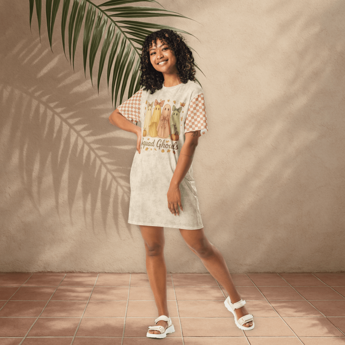 Woman wearing a ghost checkered t-shirt dress standing against a beige wall with a plant leaf.