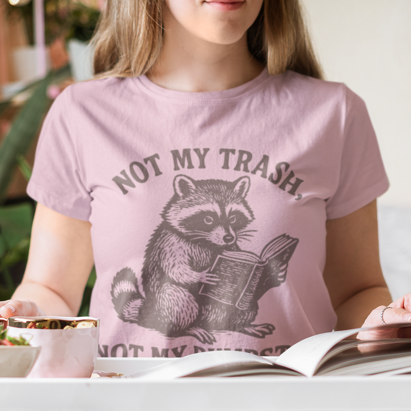 Person wearing a pink t-shirt with a raccoon graphic and 'Not My Trash' text, sitting at a table with food and an open book.