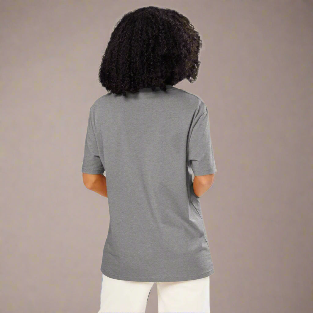 A woman with voluminous curly hair is shown from the back, wearing a plain gray short-sleeve t-shirt and white pants. She stands in front of a neutral studio background with her hands casually placed in her pockets.