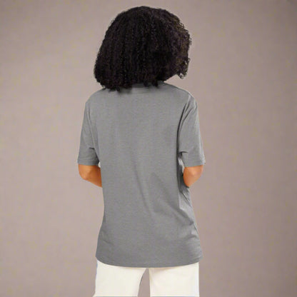 A woman with voluminous curly hair is shown from the back, wearing a plain gray short-sleeve t-shirt and white pants. She stands in front of a neutral studio background with her hands casually placed in her pockets.