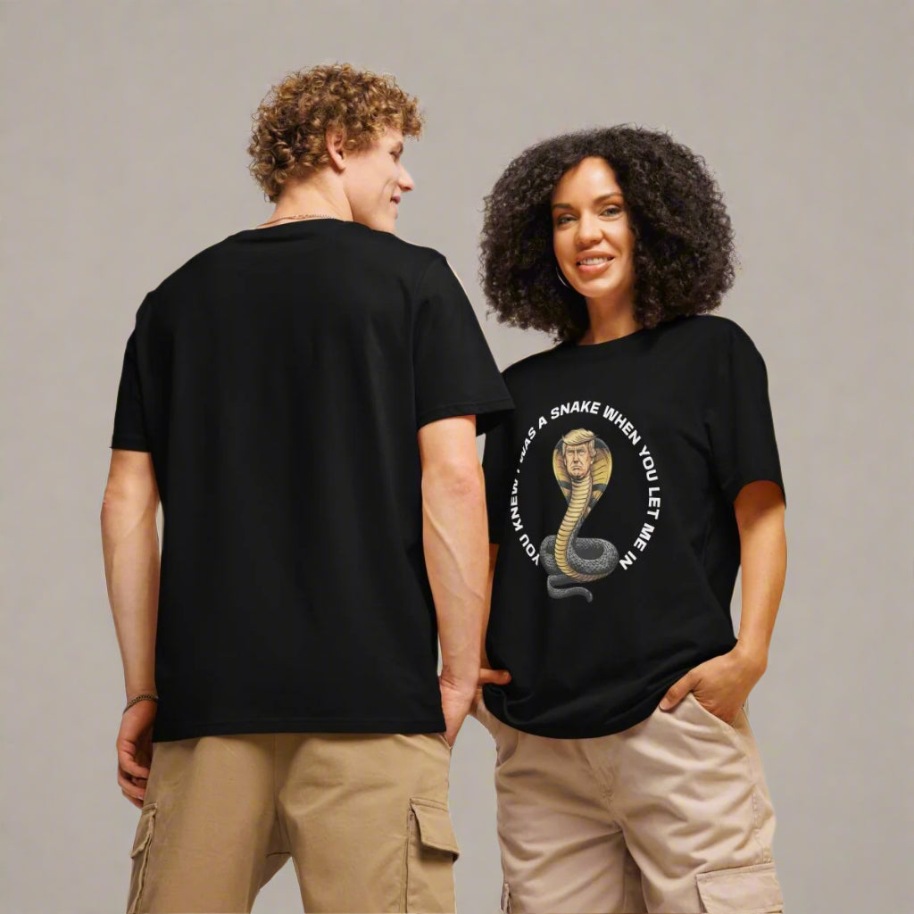A man and woman model black t-shirts with a bold political graphic. The woman faces forward, hands in her cargo shorts, smiling, while the man stands with his back to the camera. Her shirt displays an illustration of a coiled snake with Donald Trump’s face, surrounded by white text that reads: “YOU KNEW I WAS A SNAKE WHEN YOU LET ME IN.” Both wear casual neutral bottoms against a plain gray backdrop.