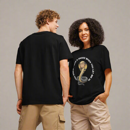 A man and woman model black t-shirts with a bold political graphic. The woman faces forward, hands in her cargo shorts, smiling, while the man stands with his back to the camera. Her shirt displays an illustration of a coiled snake with Donald Trump’s face, surrounded by white text that reads: “YOU KNEW I WAS A SNAKE WHEN YOU LET ME IN.” Both wear casual neutral bottoms against a plain gray backdrop.