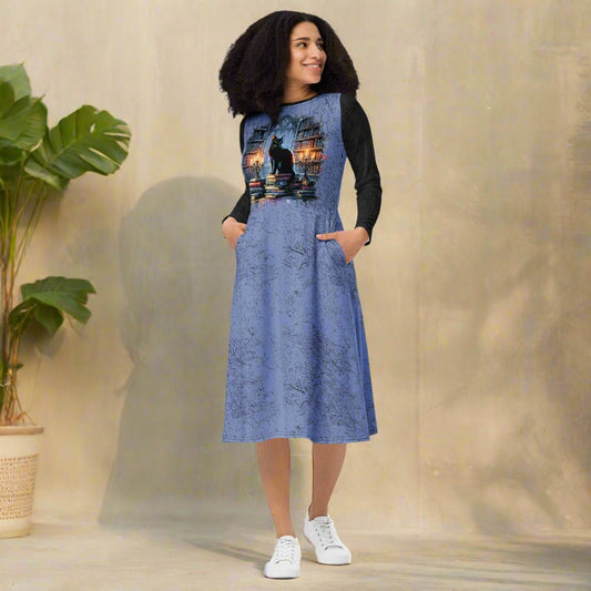 Woman wearing a blue dress with a black cat design, standing indoors next to a plant.